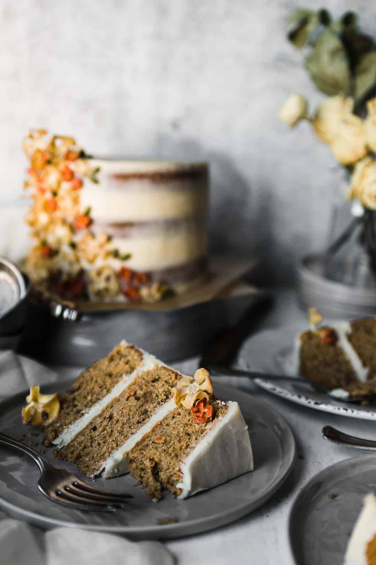 A slice of carrot spice cake on a plate with a fork and one missing bite. The whole cake is in the background and is surrounded by other plates and slices