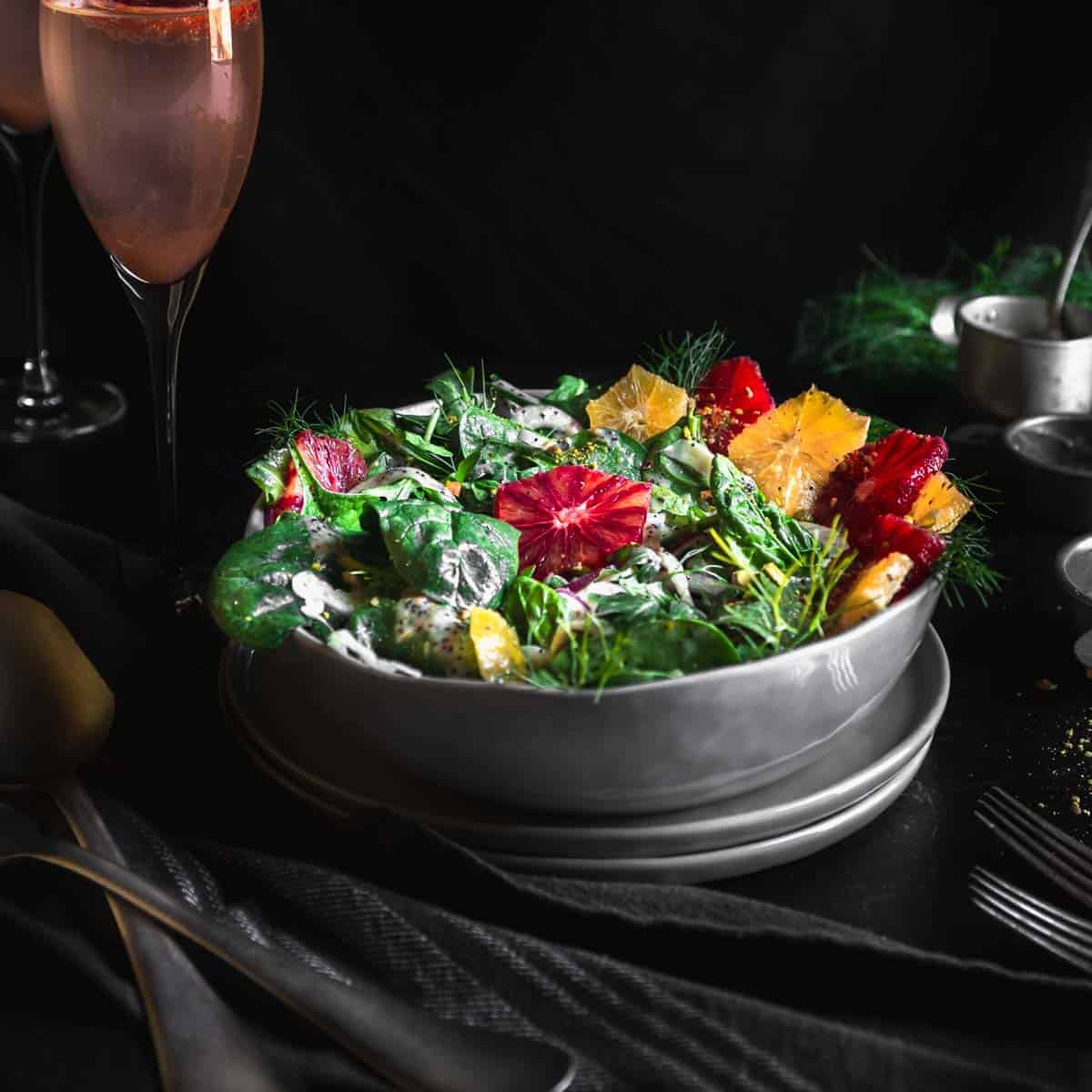 Side view of blood orange salad with poppyseed dressing in a large grey bowl. Next to the salad is forks, serving utensils, pink cocktails and a few small tin cups