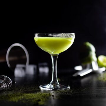 A Matcha Margarita in a matcha salted coup glass. Matcha powder, ice and squeezed limes surround the glass.