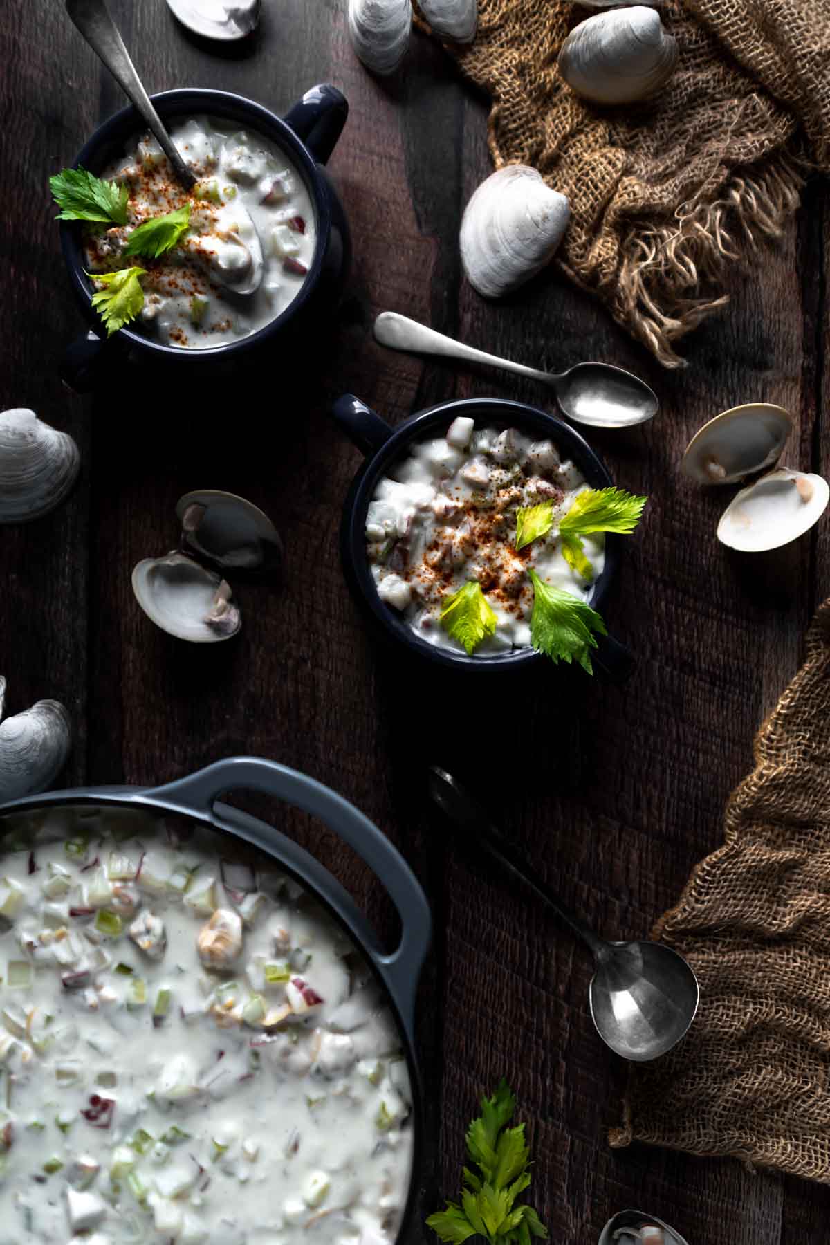 Flat Lay Photo of Chowder in a dutch oven and two bowls surrounded by Littleneck Clam Shells, spoons and a burlap net 