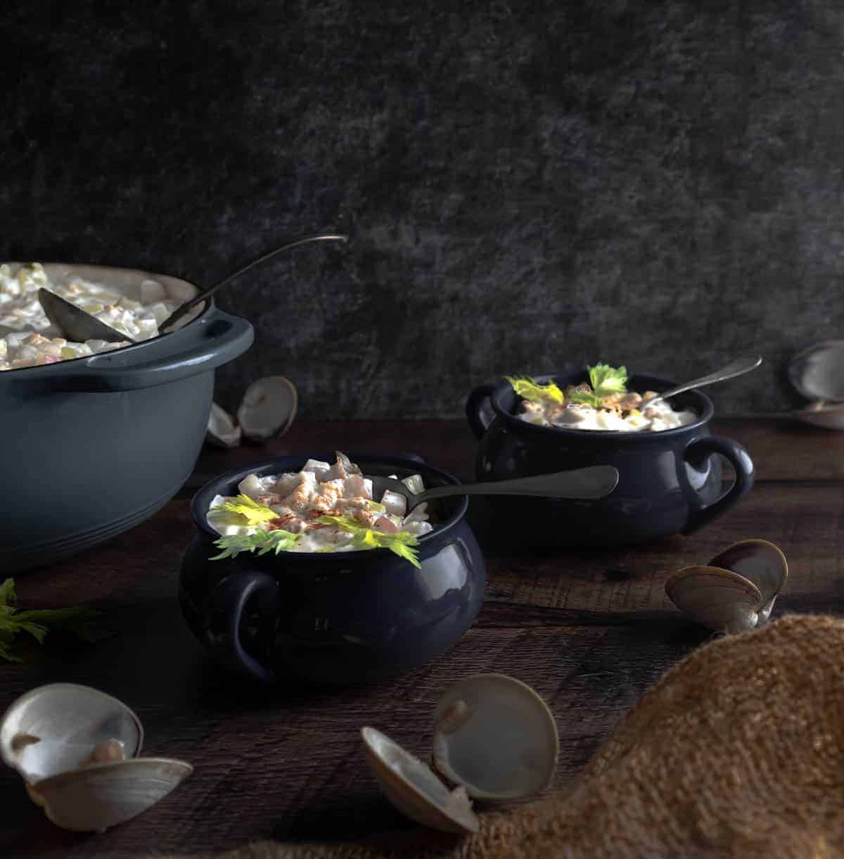 A dutch oven full of Homemade Littleneck Clam Chowder and two bowls full of chowder topped with a dusting of paprika and celery leaves