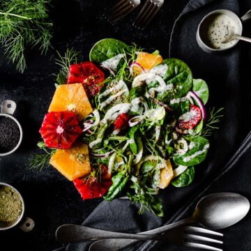 a big bowl of salad with alternating slices of orange and blood orange on one side and a drizzle of poppyseed dressing over the top. The salad is surrounded by cups of more dressing, pistachios, poppyseeds, serving utensils and fresh fennel fronds