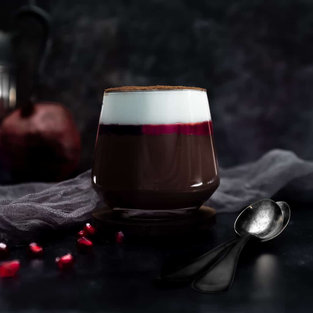 a glass filled with dark chocolate cremeaux topped with a thin layer of pomegranate gelee and a layer of whipped cream dusted in cocoa powder. The glass is next to a spoon and a sprinkle of pomegranate arils