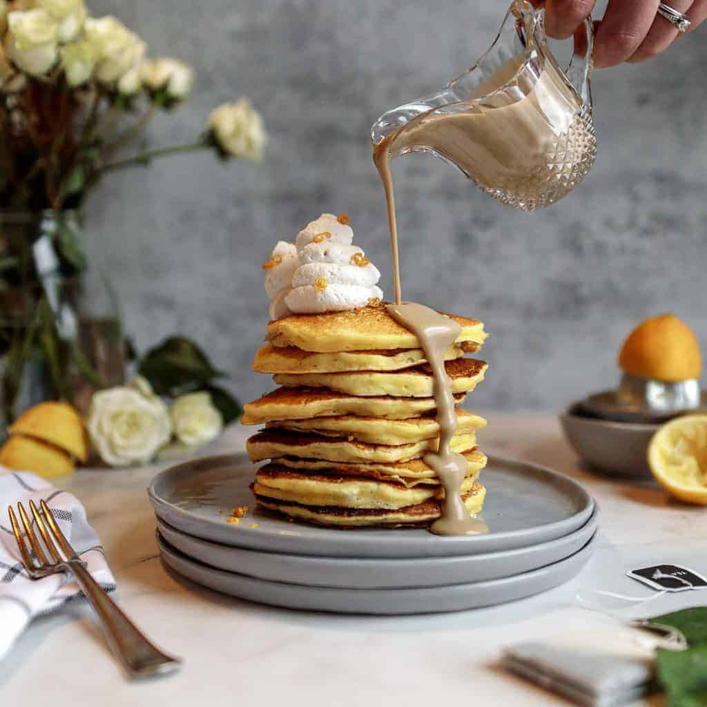 A tall stack of lemon pancakes topped with a swirl of whipped cream on a stack of grey plates surrounded by flowers and lemons. Earl grey creme anglaise is being poured over the pancakes from a crystal pourer.