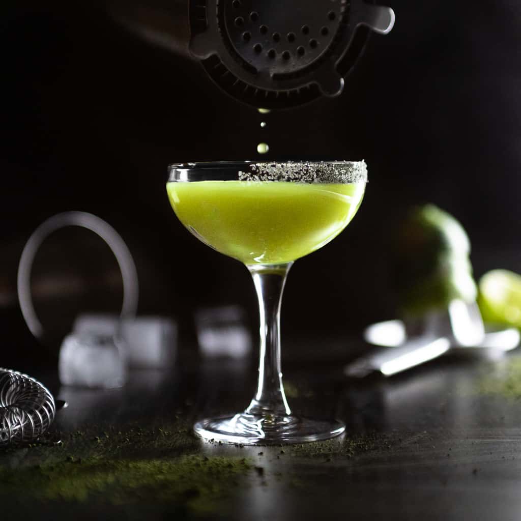 The last drops of a Matcha Margarita being poured from a cocktail shaped inso a coupe glass with a matcha salt rim