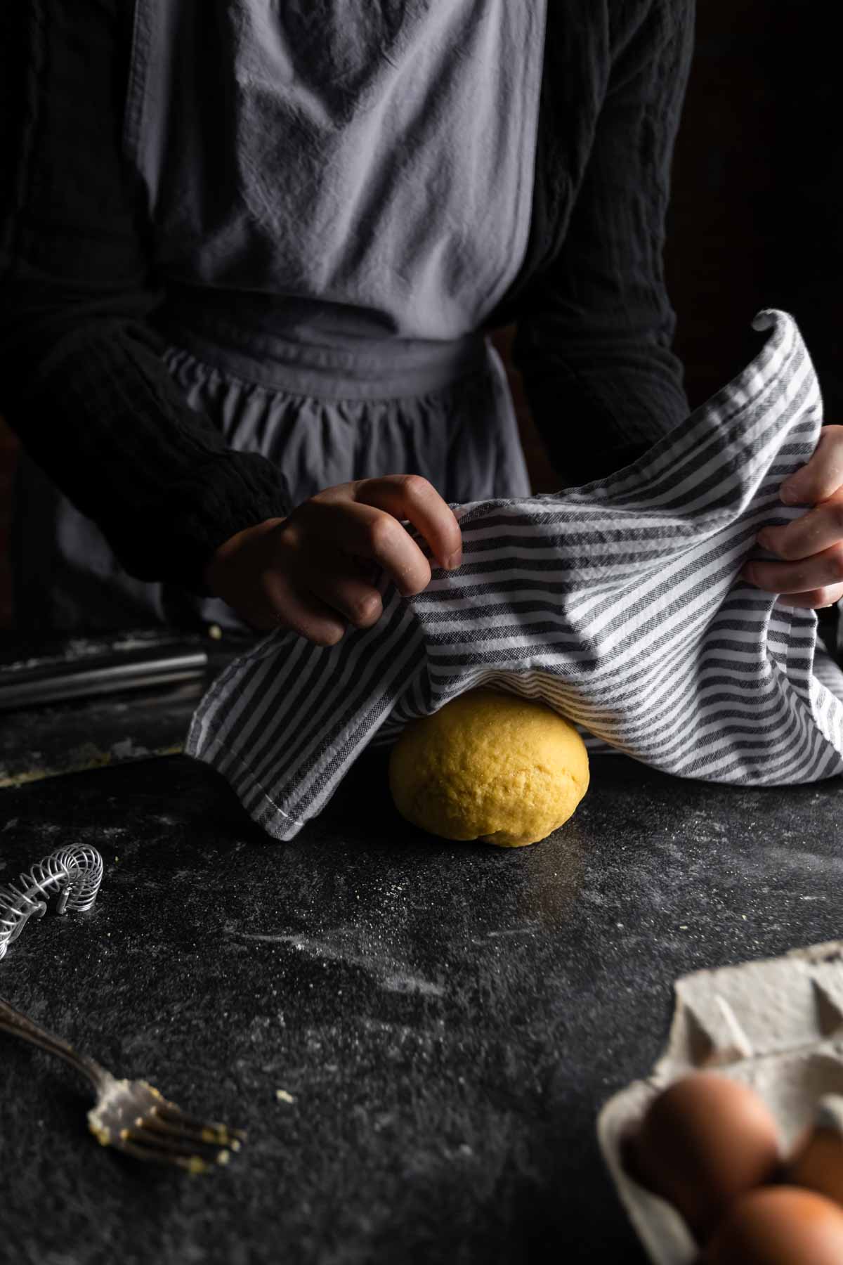 an aproned woman laying a striped kitchen towel over a ball of freshly made pasta dough