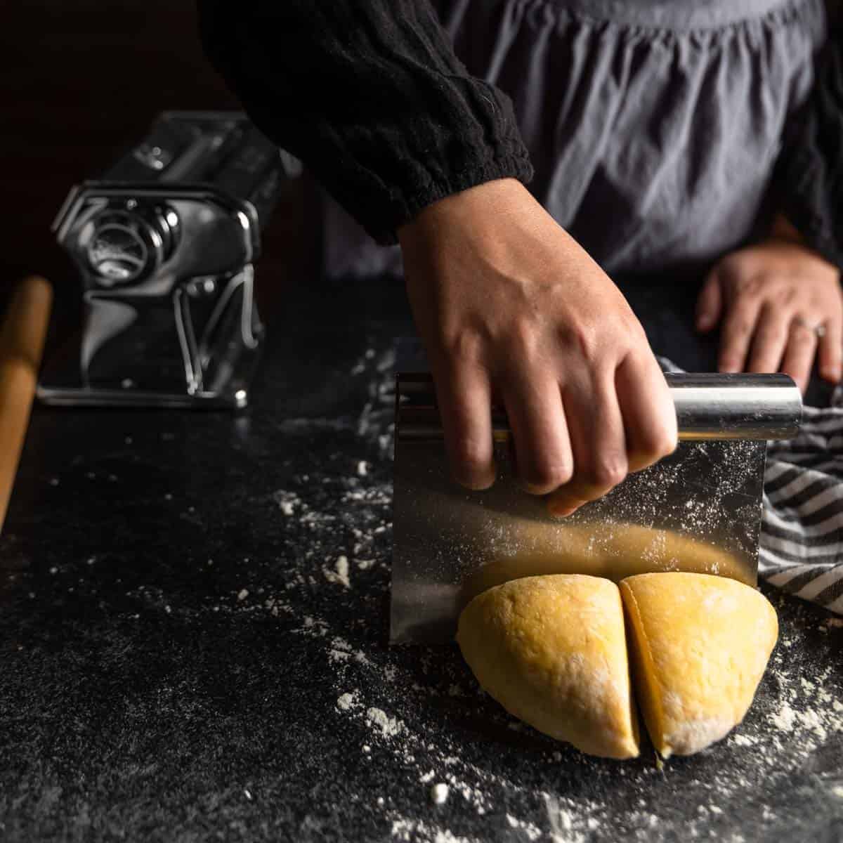 A woman cutting a ball of pasta dough into quarters with a bench scraper.