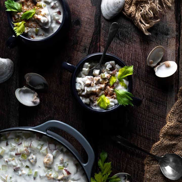 An overhead shot of two bowls of littleneck clam chowder next to clam shells, a burlap net and a blue dutch oven of more chowder