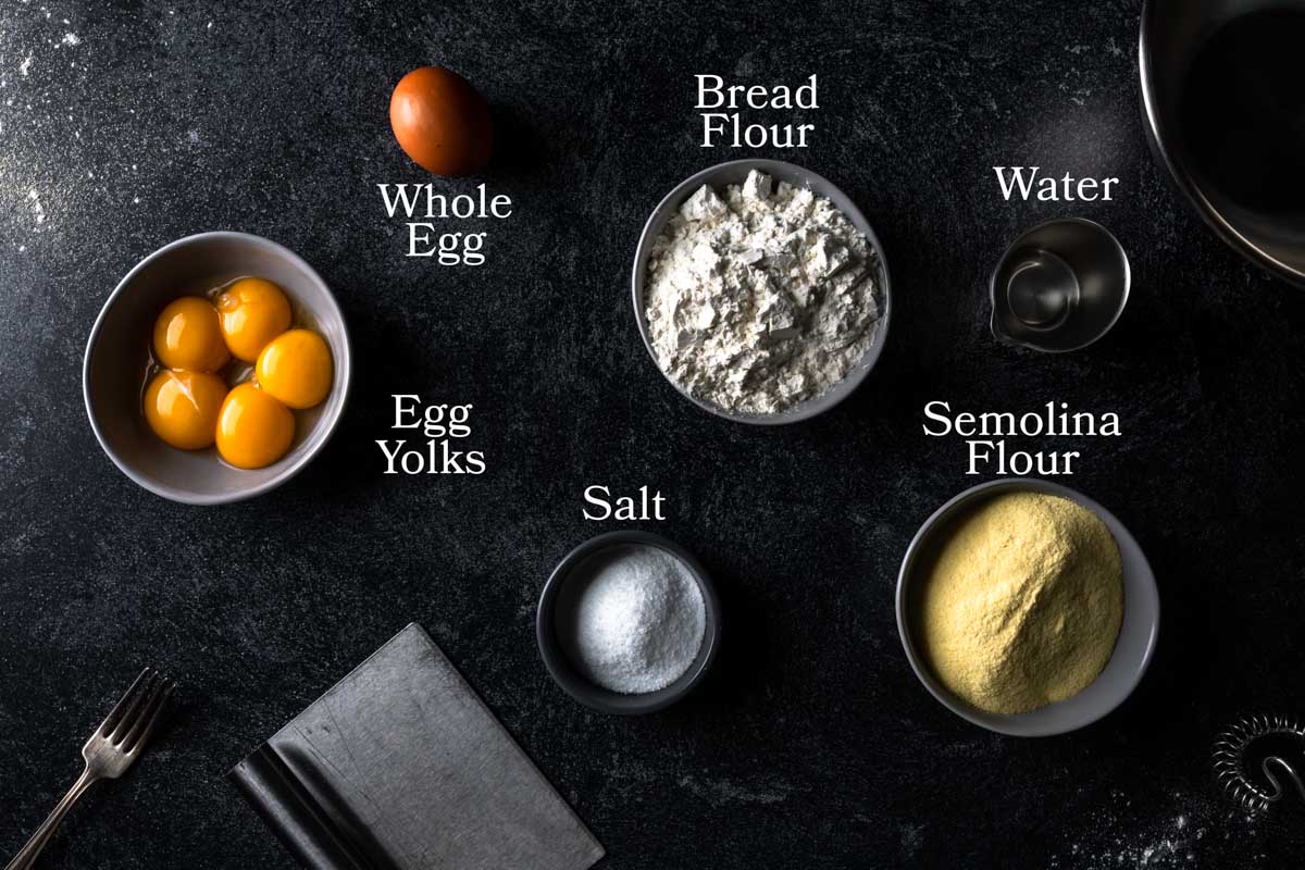 a picture of ingredients needed to make semolina pasta dough. In small bowls there are egg yolks, bread flour, semolina flour, water salt and a whole egg