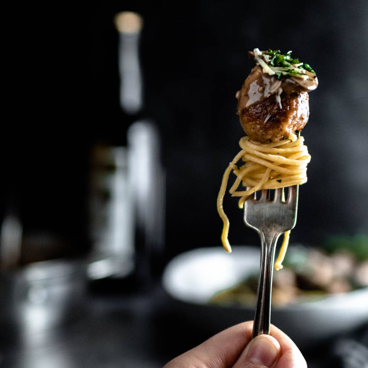 a hand holding a fork that is stabbing a chicken marsala meatball that is piled on a big bowl of homemade spaghetti