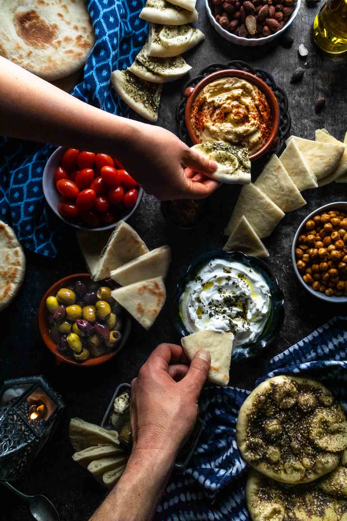 An overhead photo of two hands dipping pita bread into a colrful assortment of mezze including hummus and tzatziki dips. 