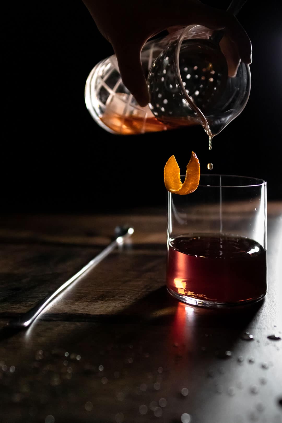 The last drops of a Cynar toronto cocktail being poured from a yari into an old fashioned glass garnished with an orange peel. The glass is next to a long bar spoon and droplets of water on the table.