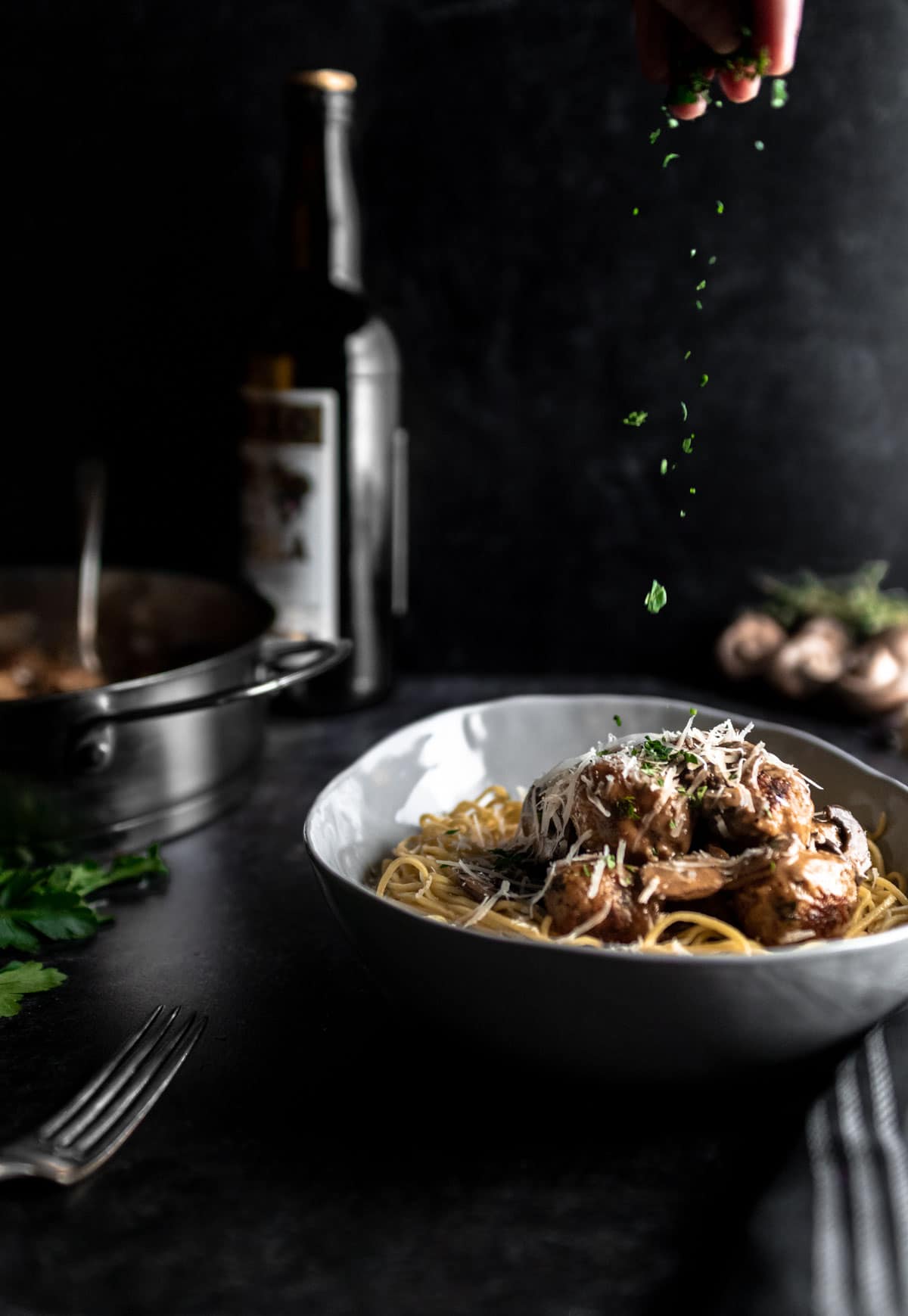 A sprinkle of fresh chopped parsley falling over a bowl of spaghetti and Chicken Marsala Meatballs with parmesan. A pan of meatballs and bottle of marsala wine is in the background