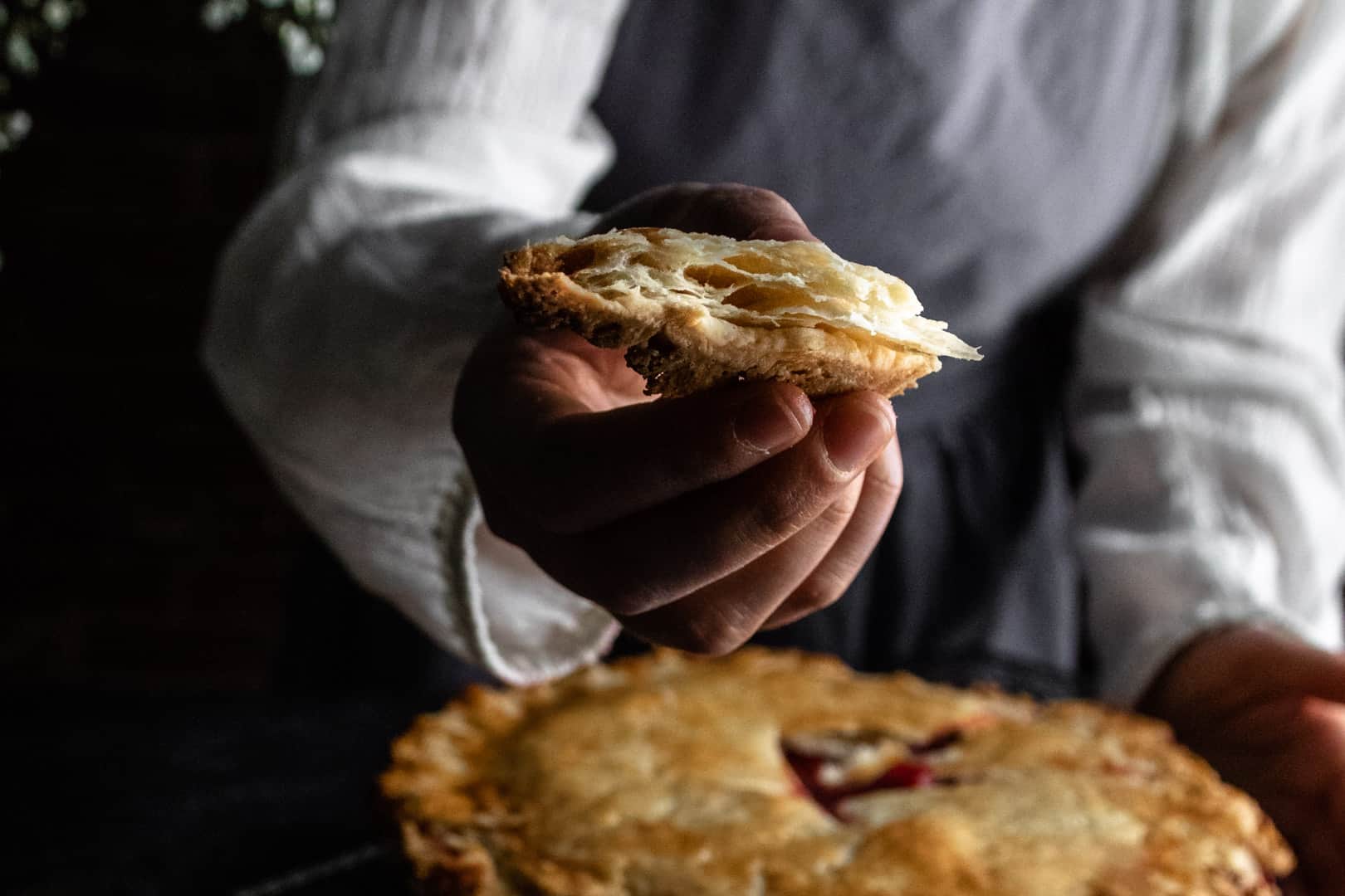 A woman holding out a close-up, super flaky piece of baked pie dough over a fresh-baked pie