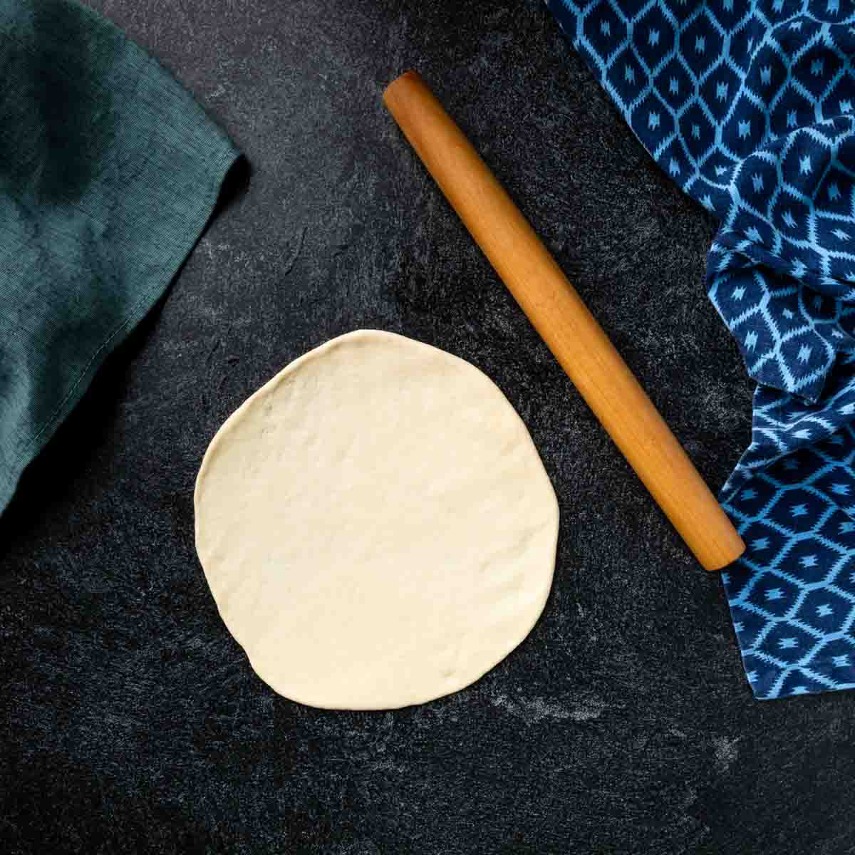 A round of pita dough rolled thin next to a small rolling pin. 