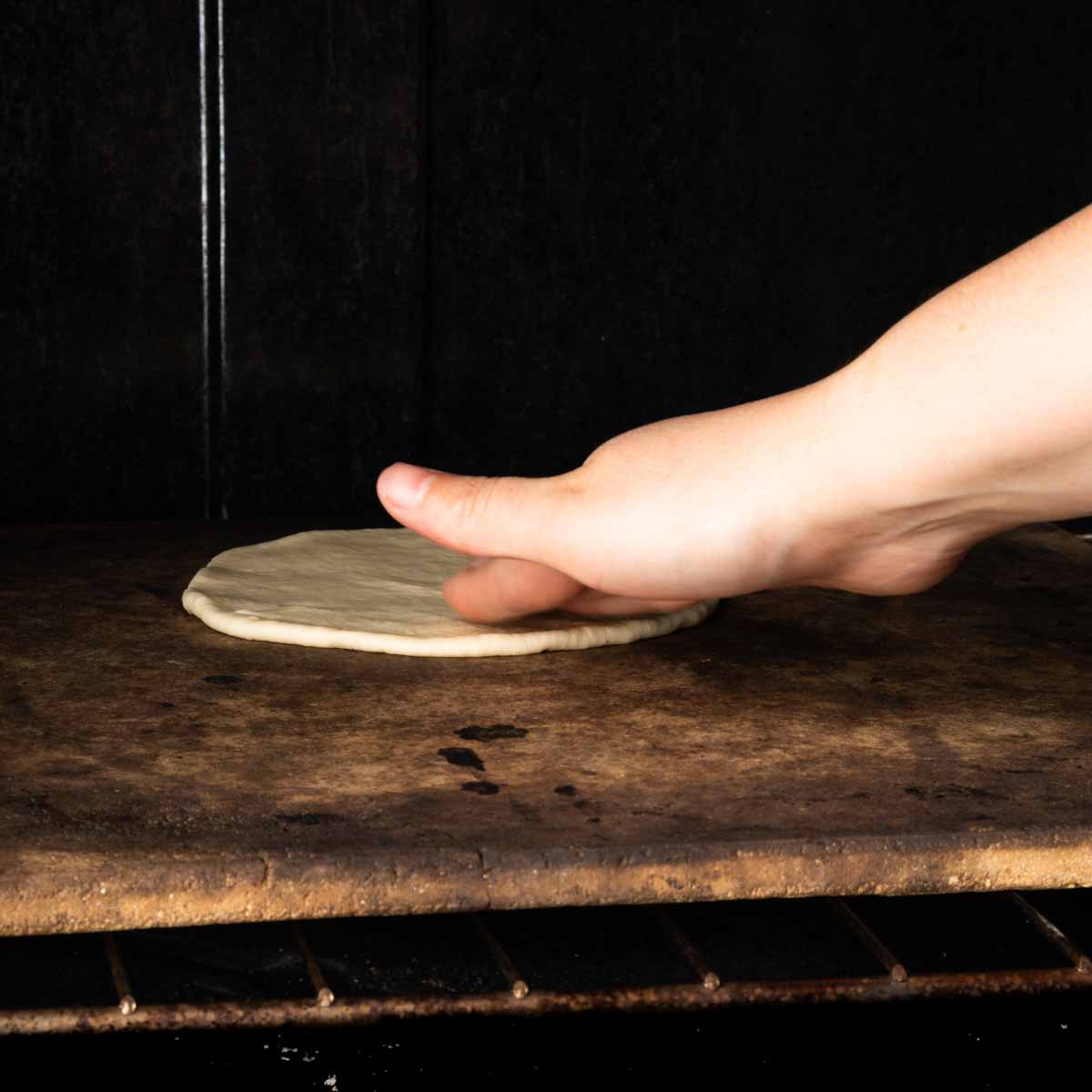 A hand placing a round of pita bread on a hot baking stone in the oven.