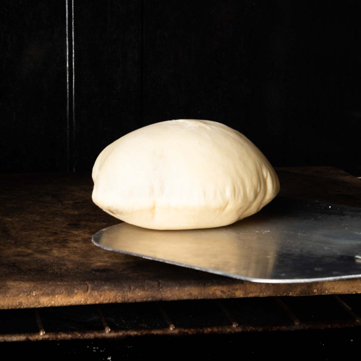 A fully puffed pita pocket being taken out of the oven on a baker's peel. 