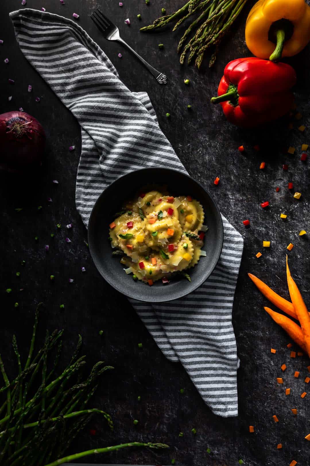 A bowl of Funfetti Ravioli Primavera on a striped linen surrounded by whole and diced red and orange bell peppers, carrots, green asparagus, red onion and a fork. 