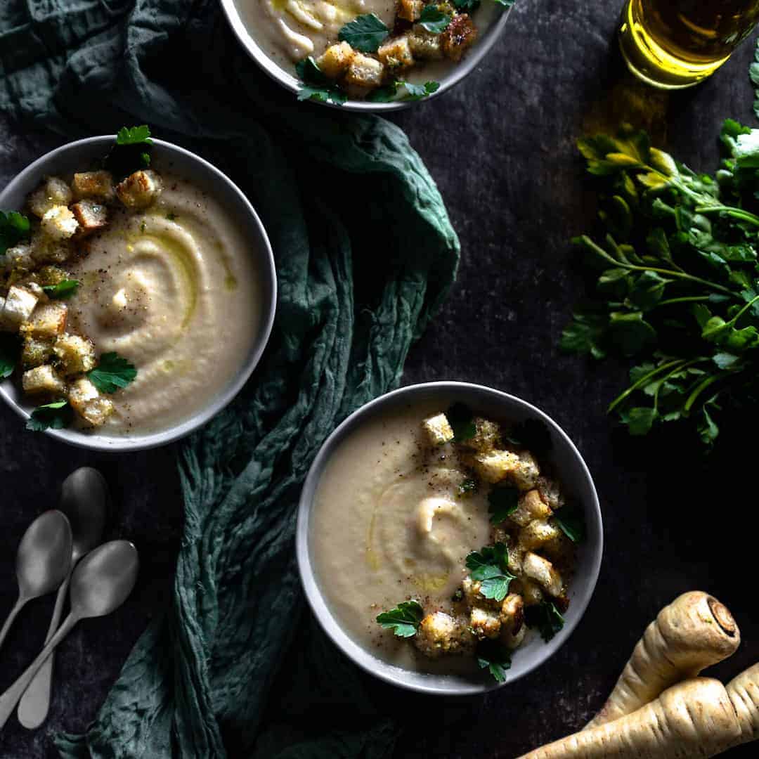 three bowls of roasted parsnip celeriac soup topped with Gremolata Croutons and staged next to spoons, a green linen, parsley and fresh parsnips