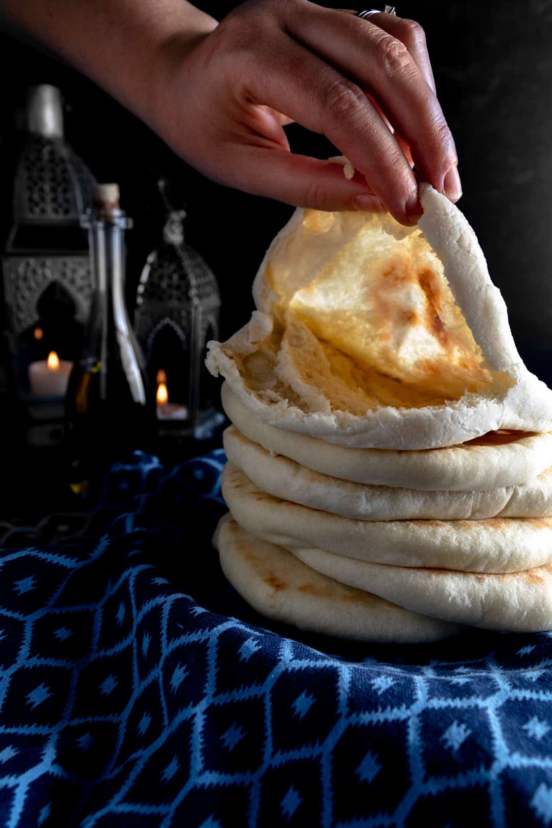 A stack of freshly baked pita bread on blue linen with the top one ripped and held open with a hand to reveal the perfect pocket. Candle lanterns are lit in the background. 