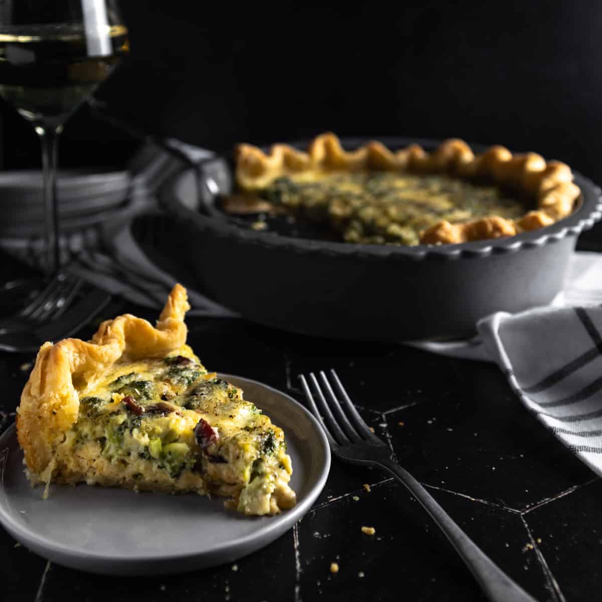 A up-close slice of Bacon Broccoli Quiche on a small plate with a fork. The whole quiche in the pie plate, a stack of small plates and a glass of white wine are in the background