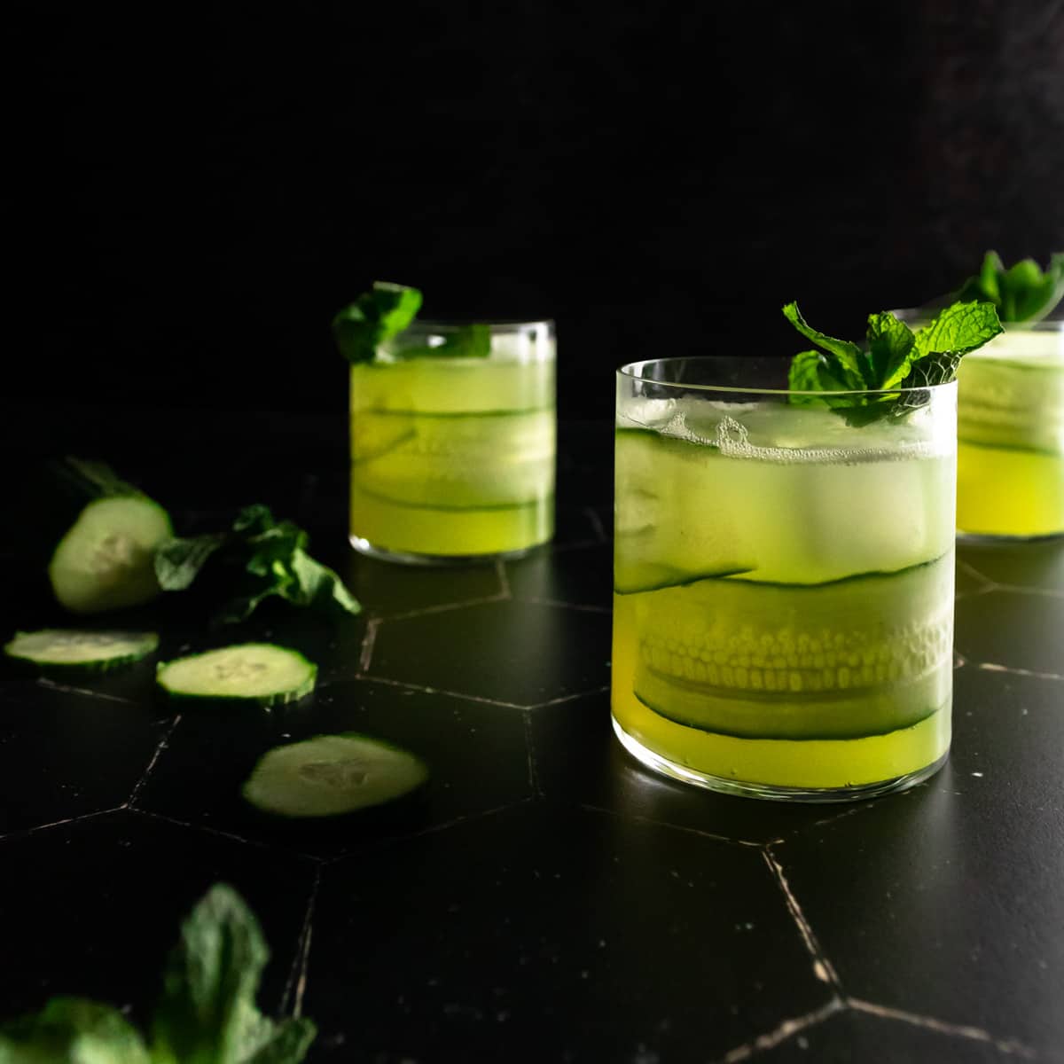 Three rocks glasses filled with cucumber mojitos garnished with a cucumber ribbon in the glass and a fresh mint sprig. A sliced cucumber and bunch of mint is in the background