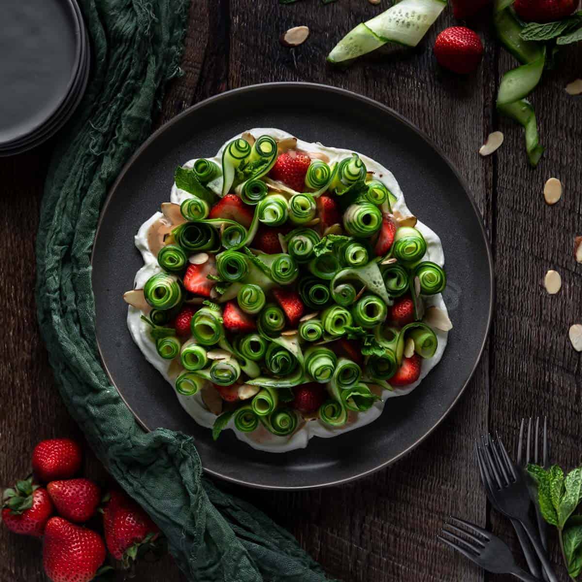 An up close shot of the cucumber strawberry salad surrounded by forks a sprinkle of toasted almond slices, strawberries and a stack of plates