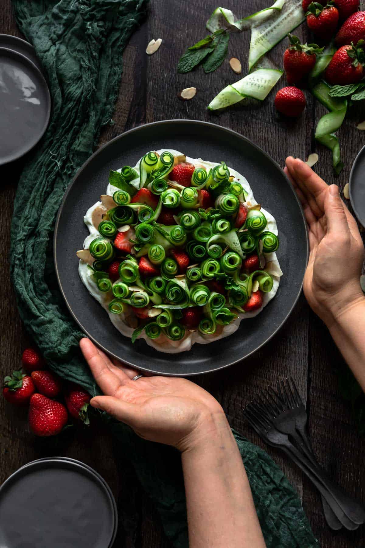 Two hands placing down a plate of curled cucumber ribbon salad with fresh strawberries and toasted almonds on a bed of whipped goat cheese. A green linen, forks, plates, and additional strawberries and slices of cucumber surround the plate. 