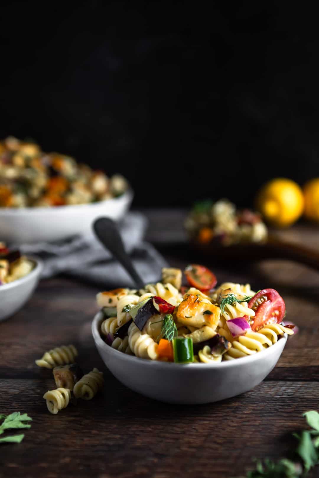 A small dish filled with Halloumi pasta salad with a fork in it and a few rotini that fell out beside the dish. A large serving bowl of the pasta salad and lemons are in the background