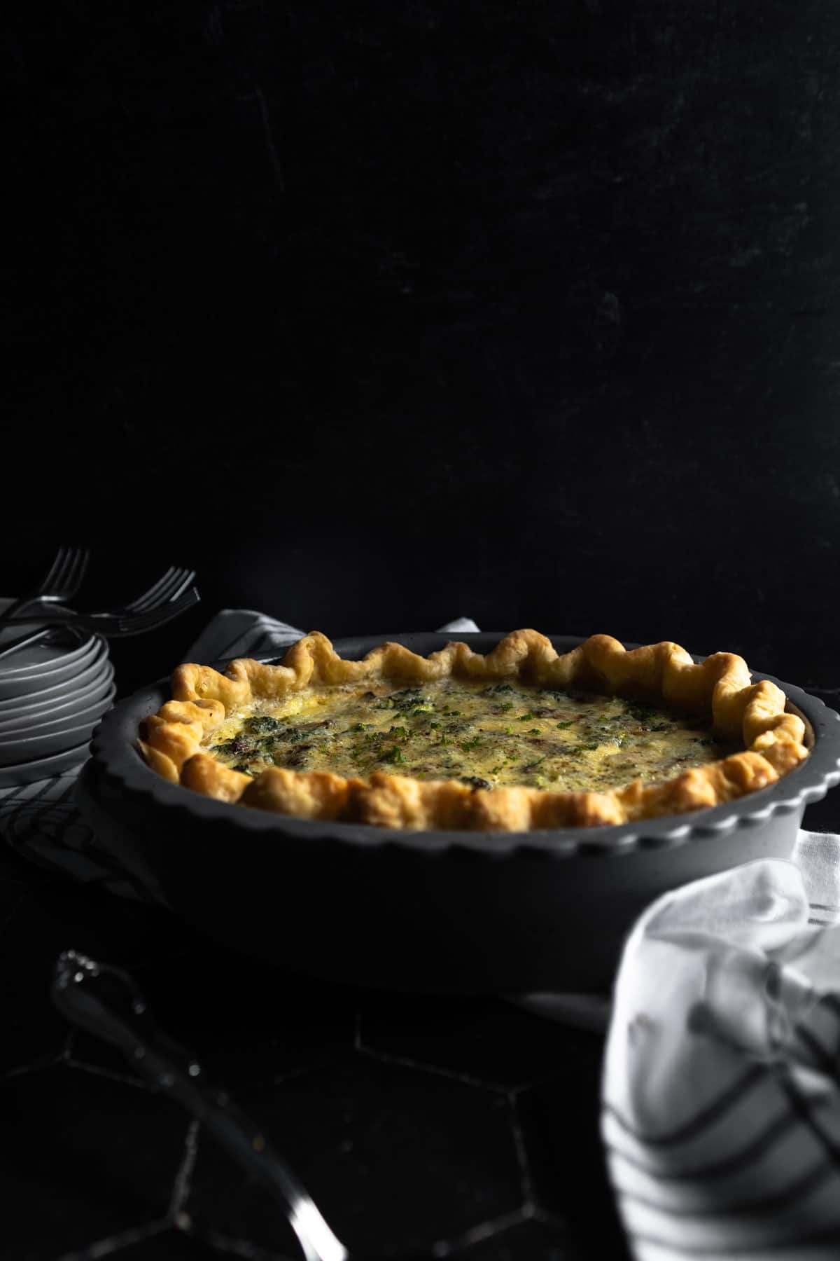 A full bacon broccoli quiche with a crimped pie edge. A stack of plates topped with forks is in the background. 