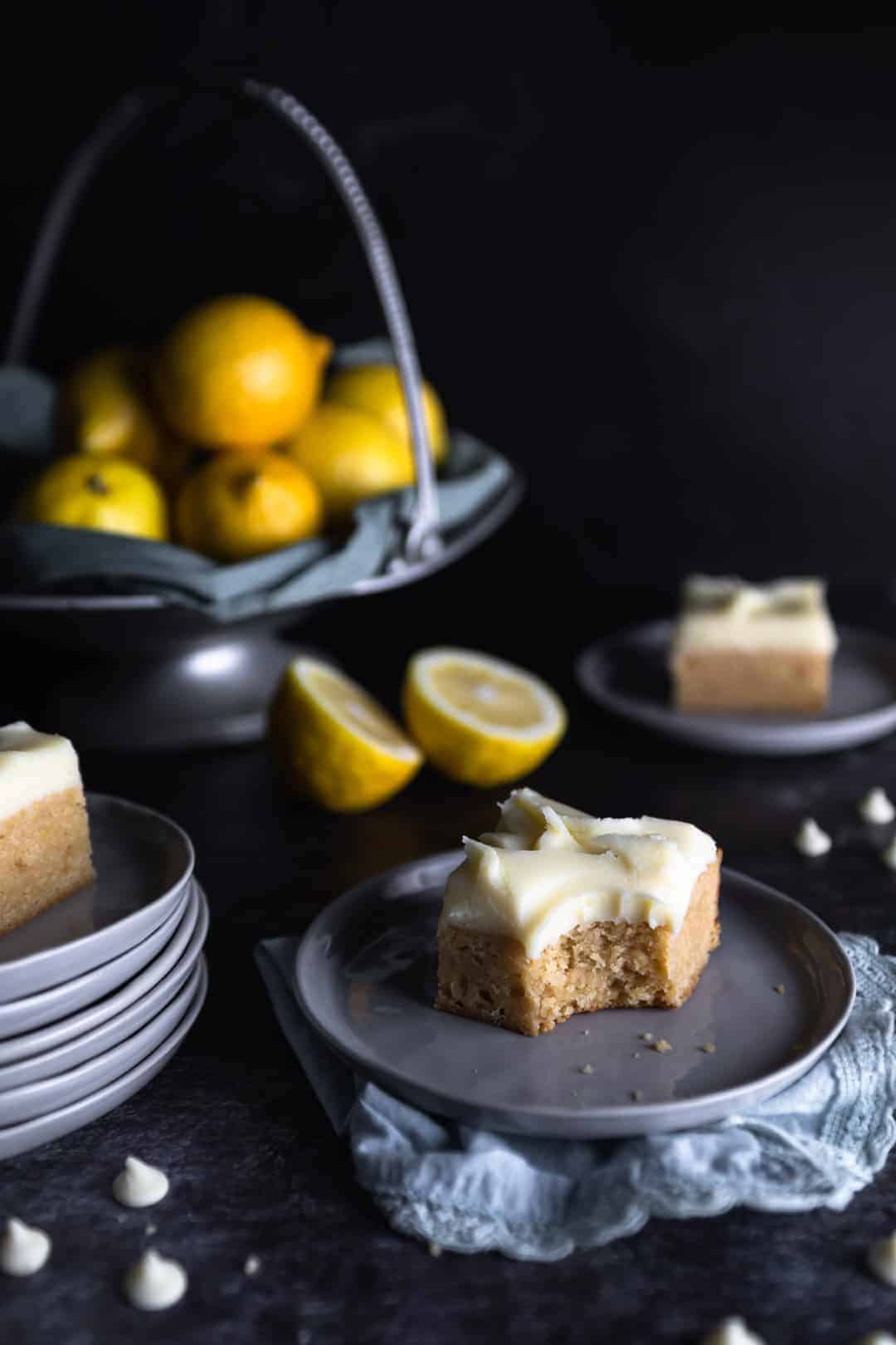 A lemon blondie with white chocolate ganache frosting on a small plate with one bite taken out. Two other blondies are on small plates nearby, white chocolate chips are scattered around and a big bowl of lemons are in the background
