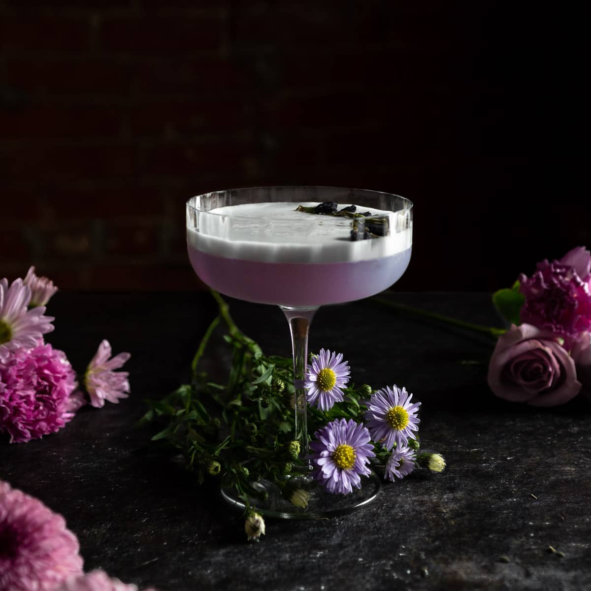 A fluted coupe glass filled with a purple Lavender Gin sour made with Empress 1908 Gin surrounded by fresh purple flowers