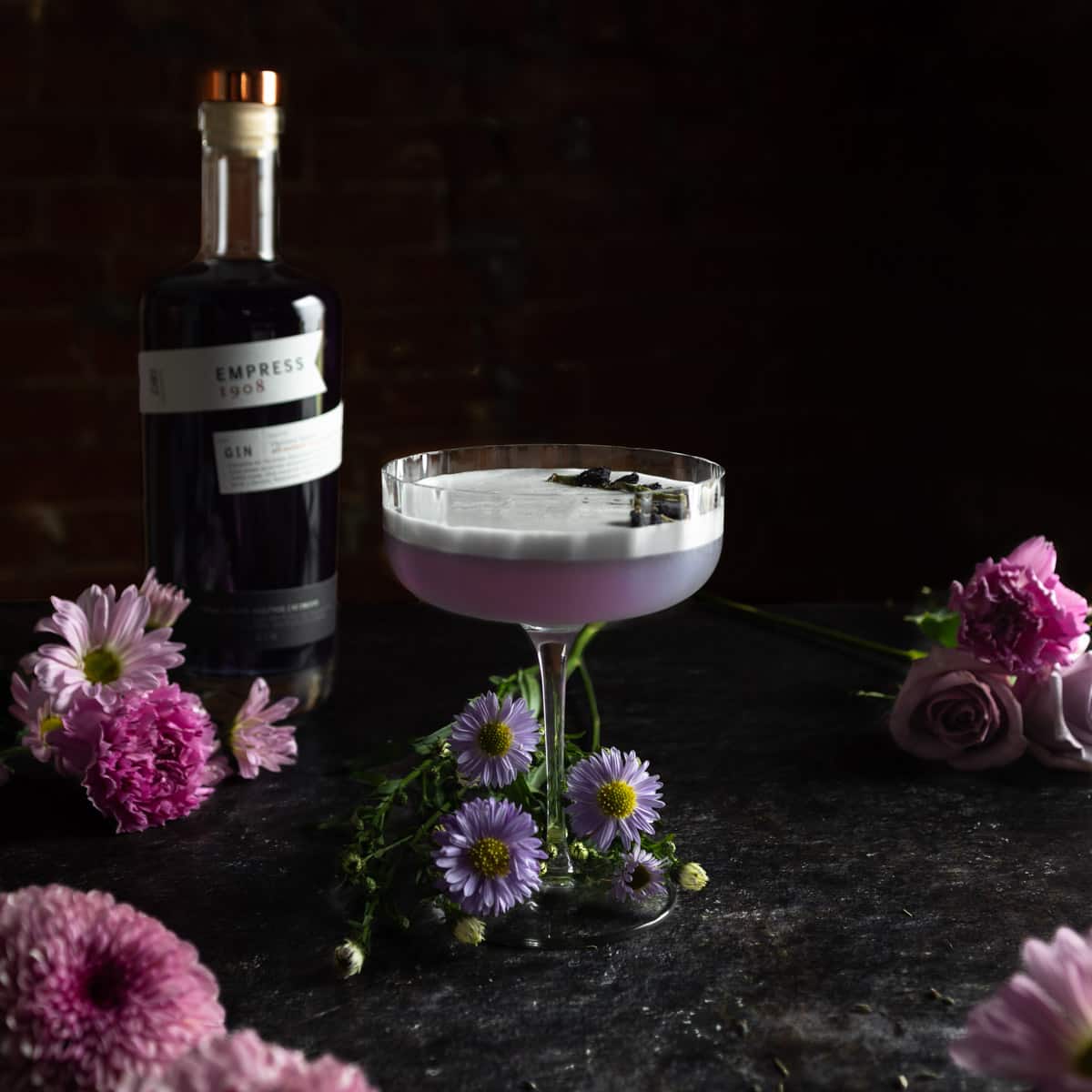 A lavender gin sour next to a bottle of Empress 1908 gin surrounded by purple flowers