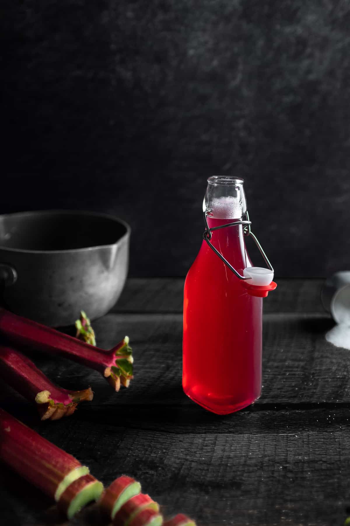 A small bottle of bright pink rhubarb syrup with a pot in the background next to s spilled cup of sugar and a stalk of chopped rhubarb