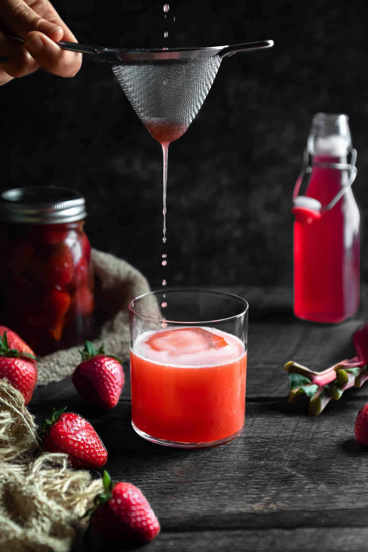 The last drops of a Strawberry Rhubarb Whiskey Sour being poured through a tea strainer into a rocks glass. A jar of strawberry infused whiskey with a burlap cloth, fresh strawberries and a bottle of rhubarb syrup are in the background.