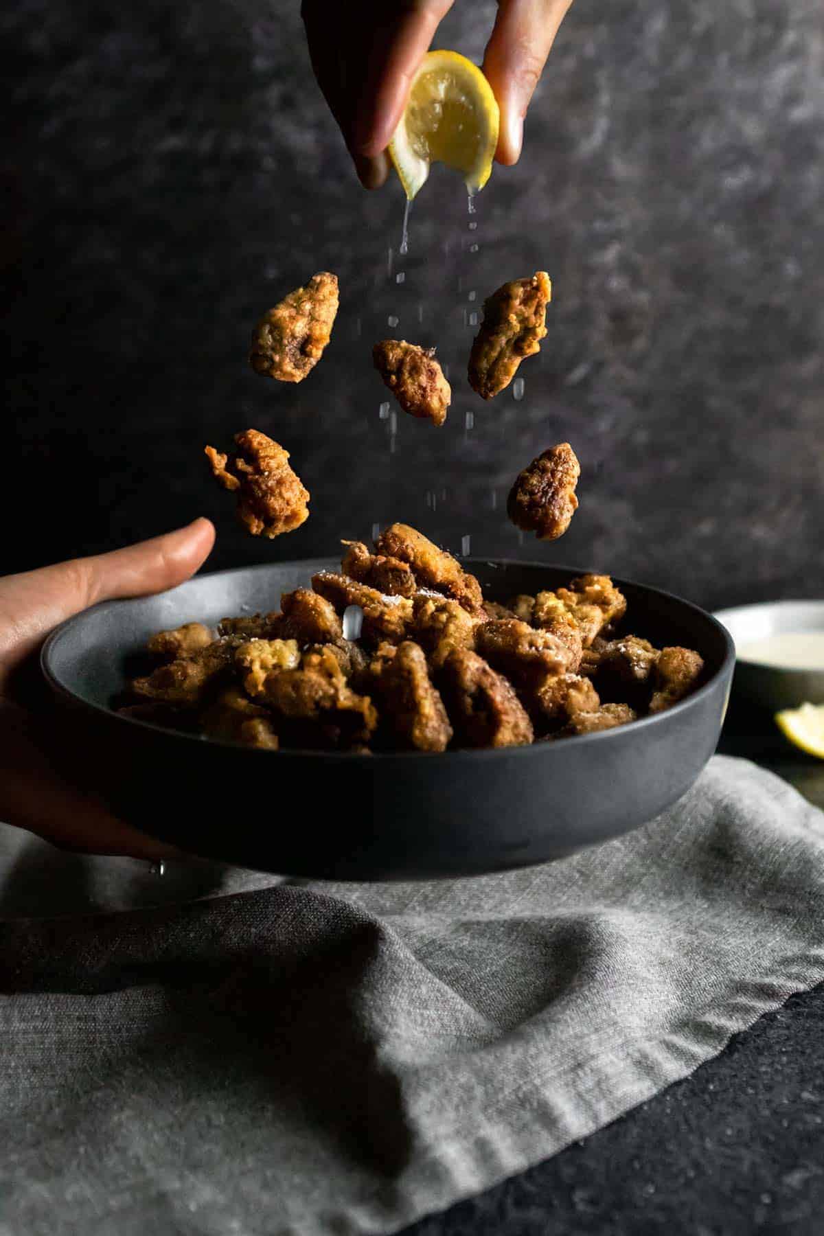 A levitation shot of fried morel mushrooms falling into a big bowl while a lemon wedge is squeezed over top