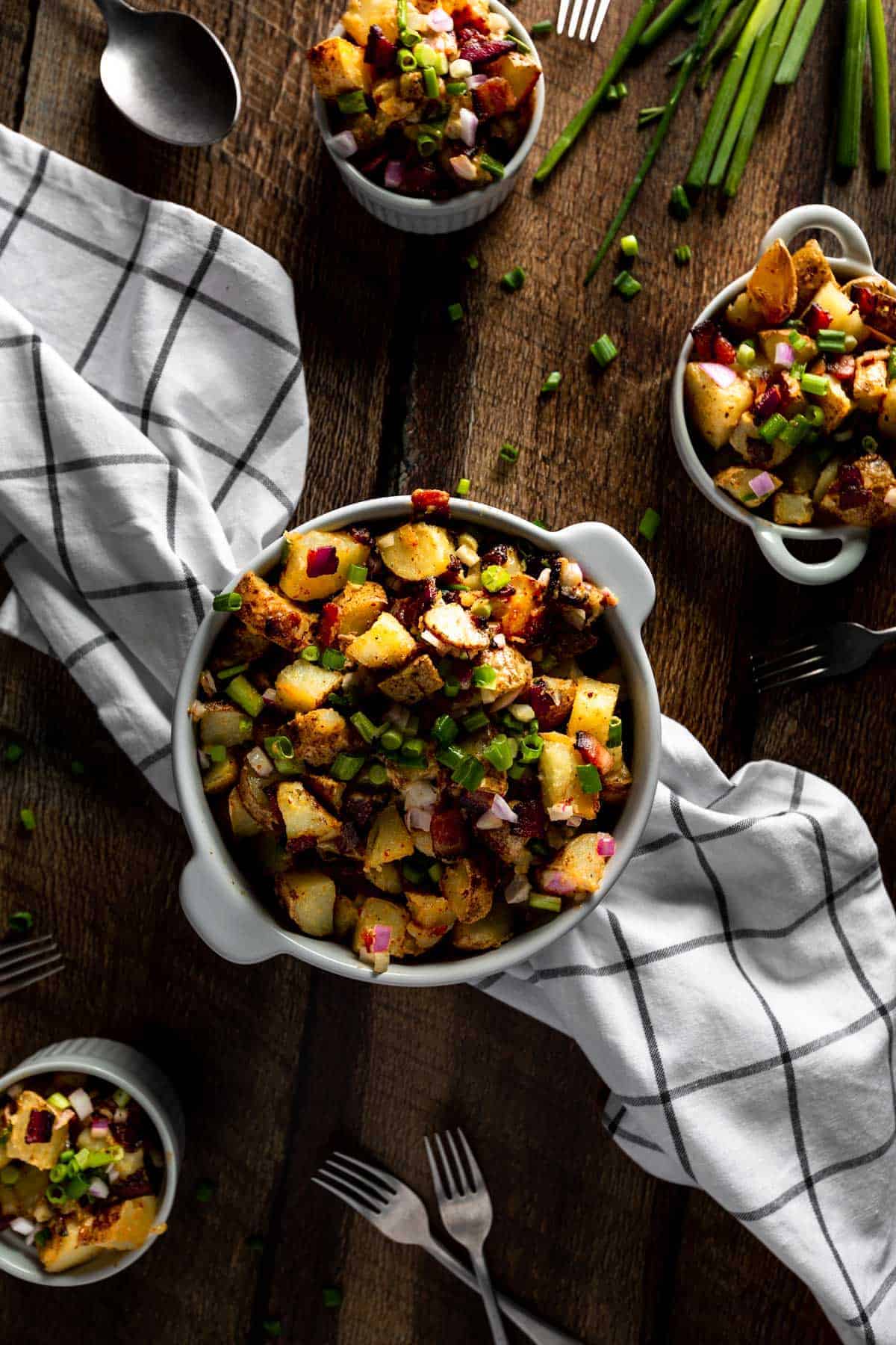 A large serving dish on a checkered linen and two small serving bowls of fried potato salad with bacon and scallion