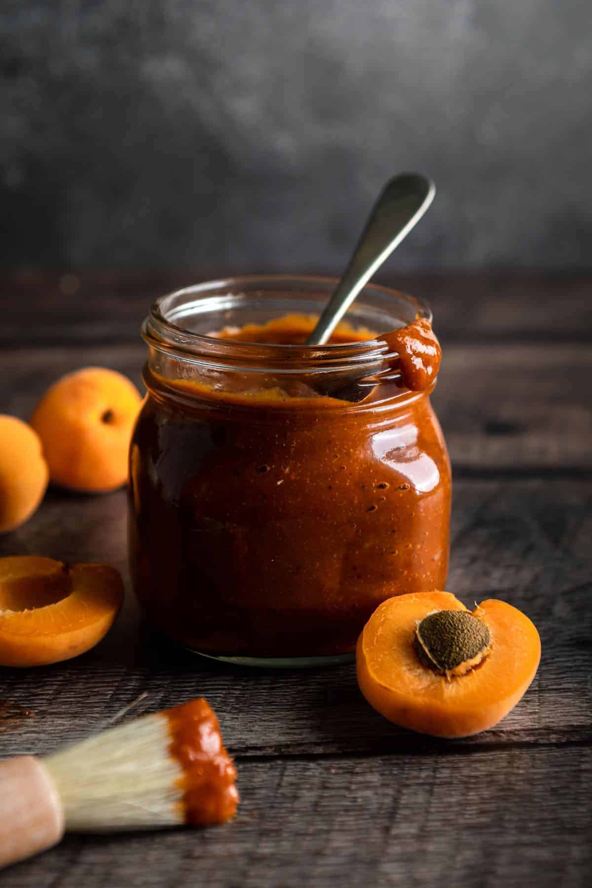 A jar of apricot bbq sauce next to a barbecue dipped basting brush and whole and half apricots