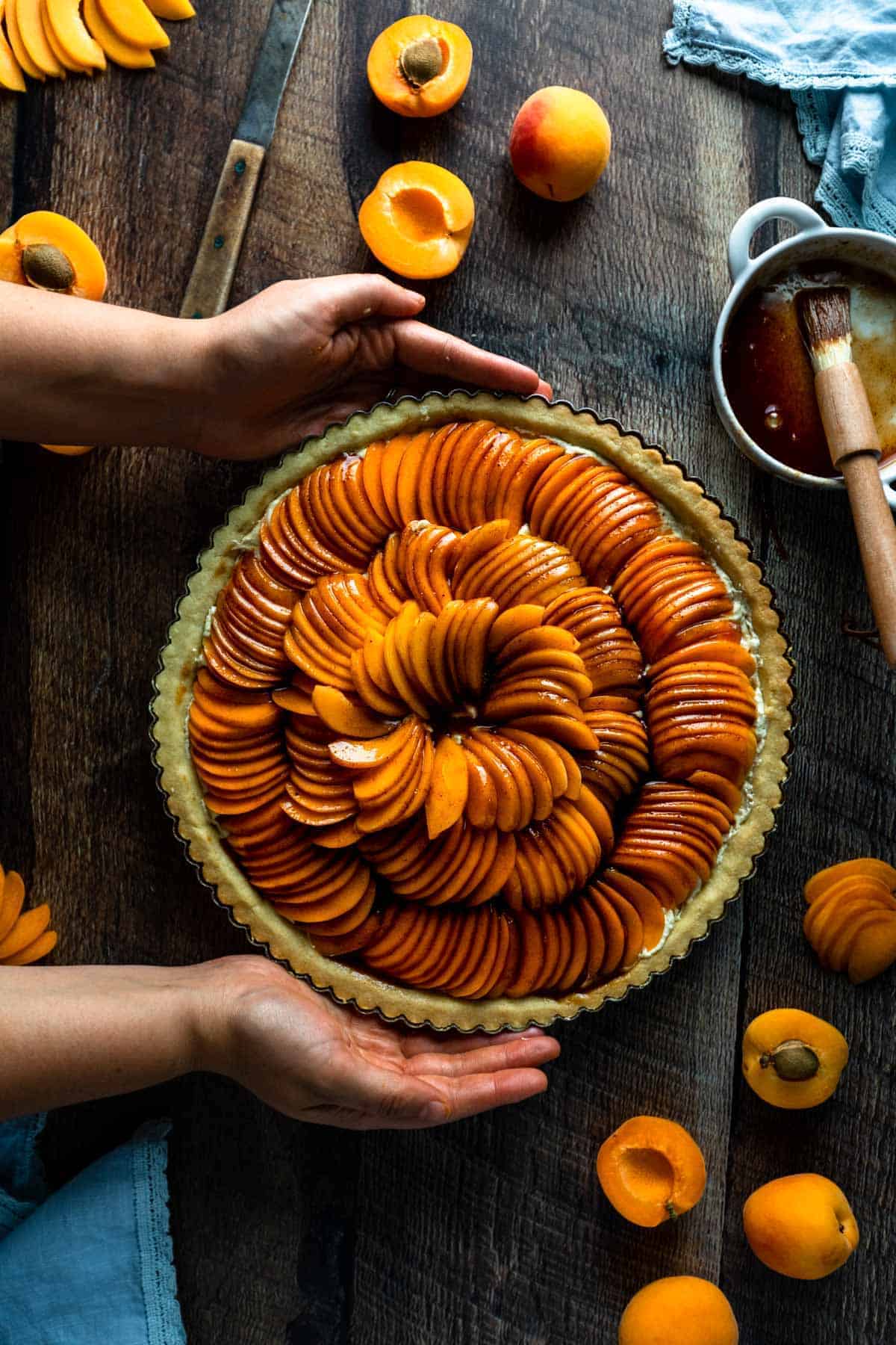 Two hands picking up an apricot tart glazed with cinnamon syrup and ready to be baked