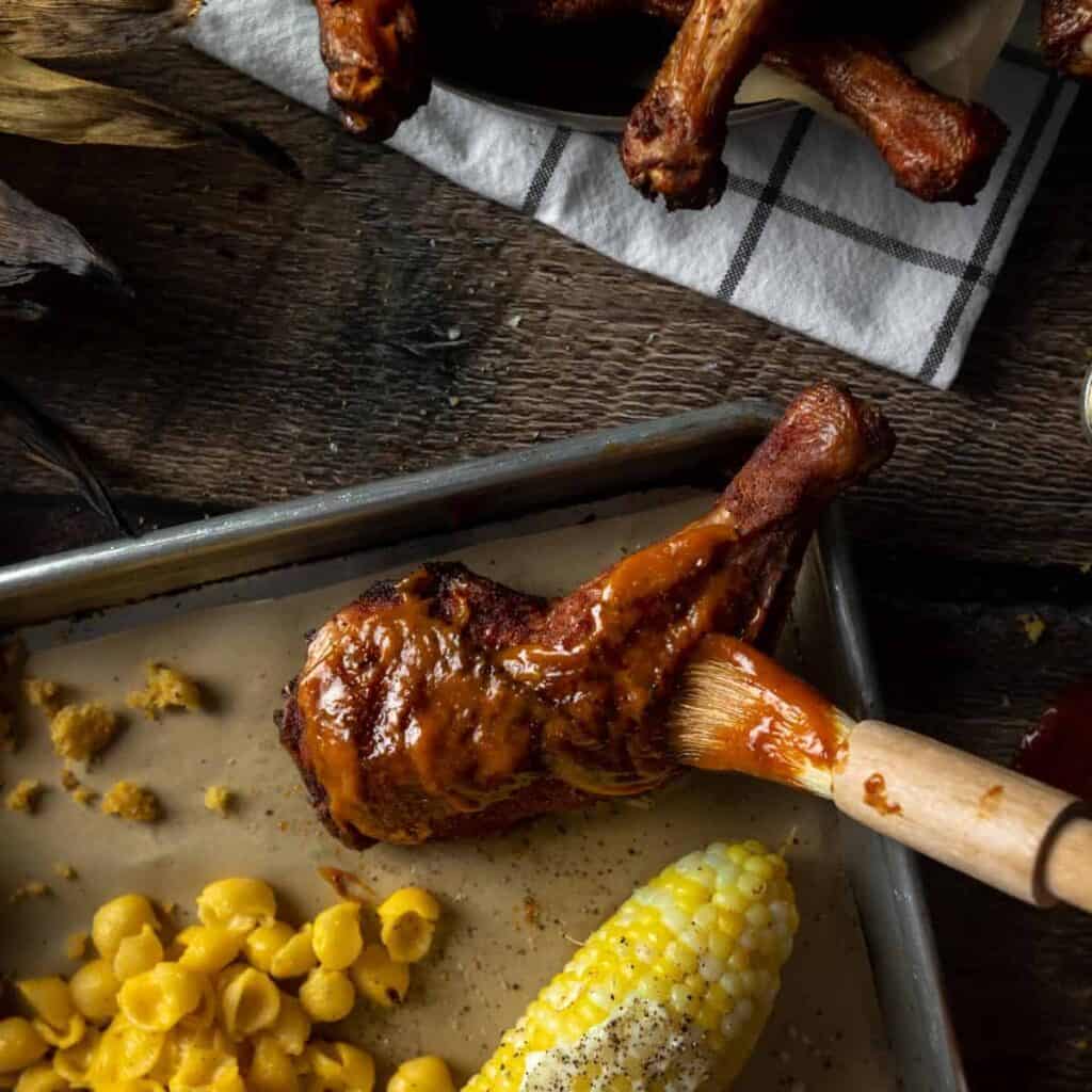 A tray of BBQ food including a smoked chicken leg being glazed with a brush of Apricot BBQ Sauce