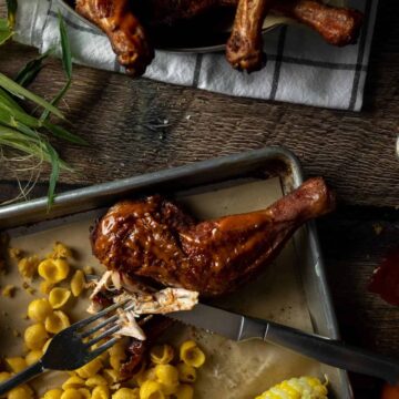 A fork and knife tearing off a piece of BBQ rubbed smoked chicken quarter on a tray next to Mac and cheese and corn on the cob