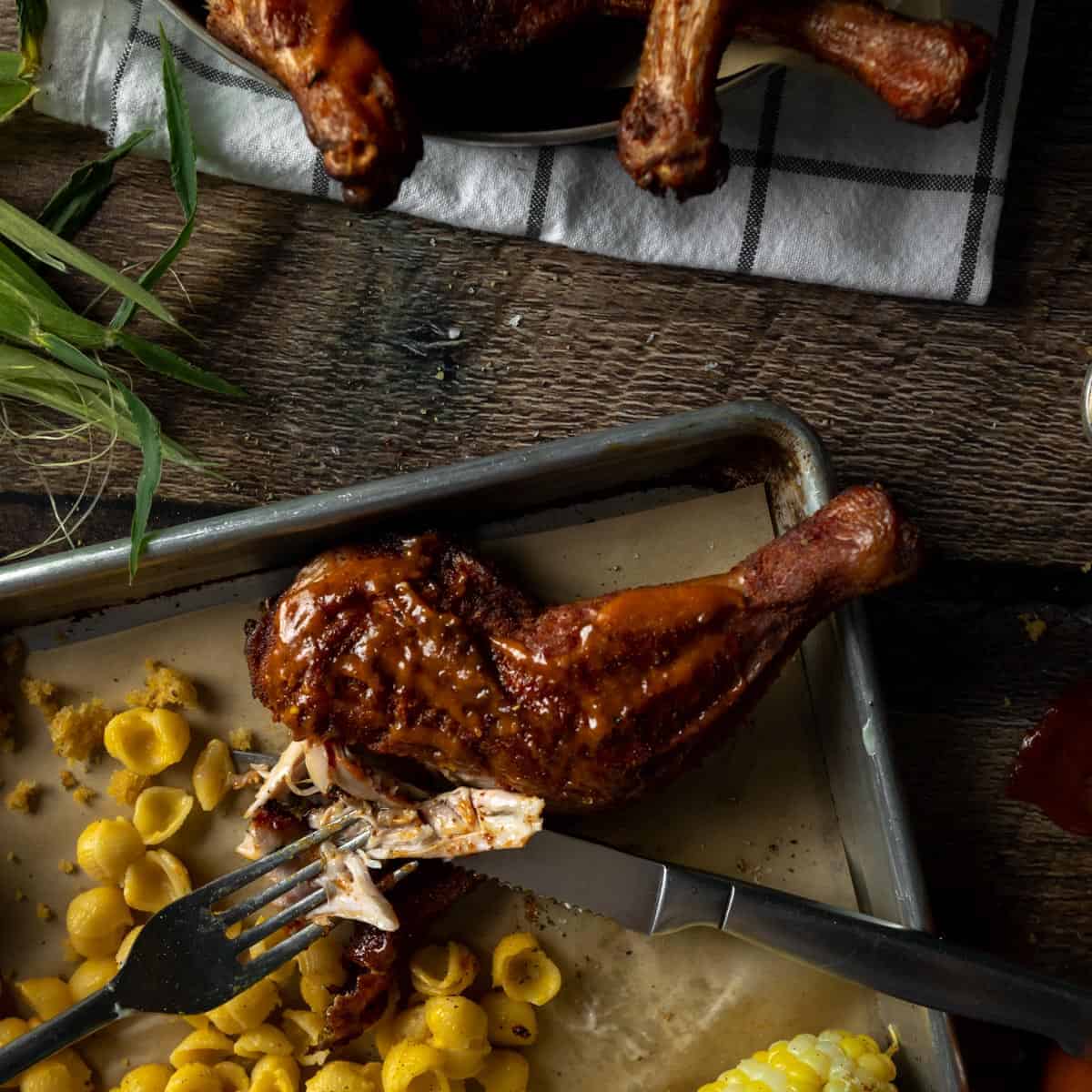 A fork and knife tearing off a piece of BBQ rubbed smoked chicken quarter on a tray next to Mac and cheese and corn on the cob