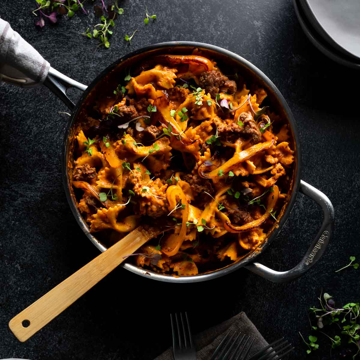 A skillet full of creamy cajun style farfalle pasta with andouille sausage that has a wooden spoon in it and a sprinkle of spicy micro greens on top