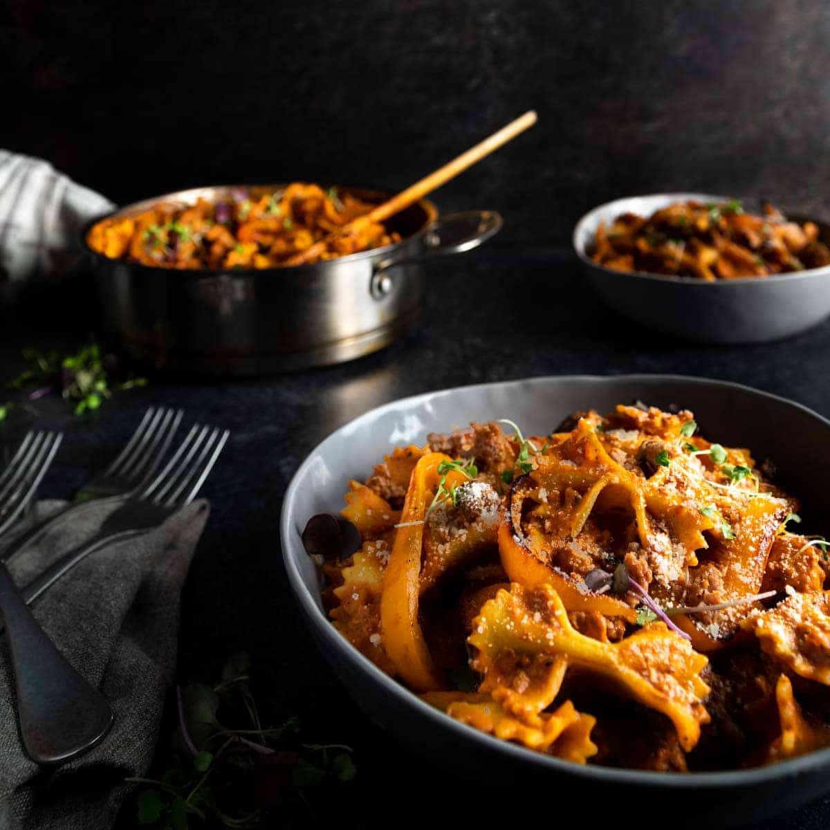 A big bowl of Cajun-style pasta with andouille sausage made with bowtie pasta and topped with a sprinkle of Parmesan and micro greens. Forks are next to the bowl and an additional bowl and skillet of the pasta are in the background.