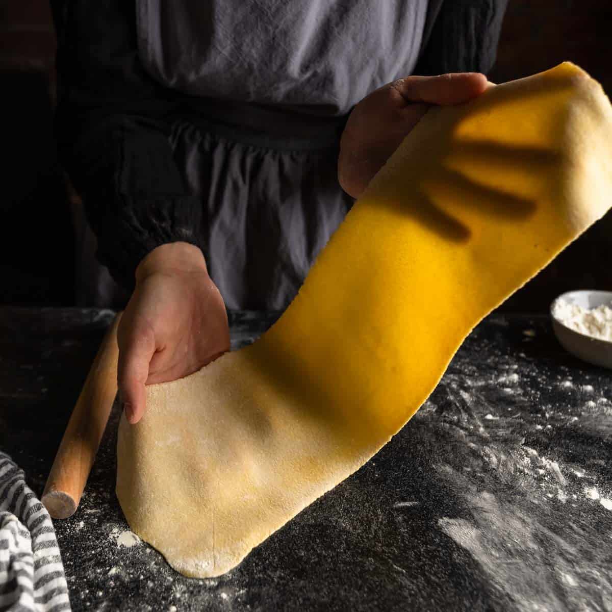 Hands holding up a translucent sheet of semolina pasta dough.