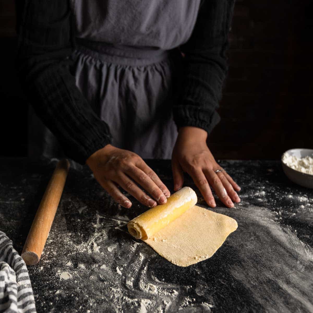 A tightly rolled spiral of sheeted pasta dough on a floured counter.