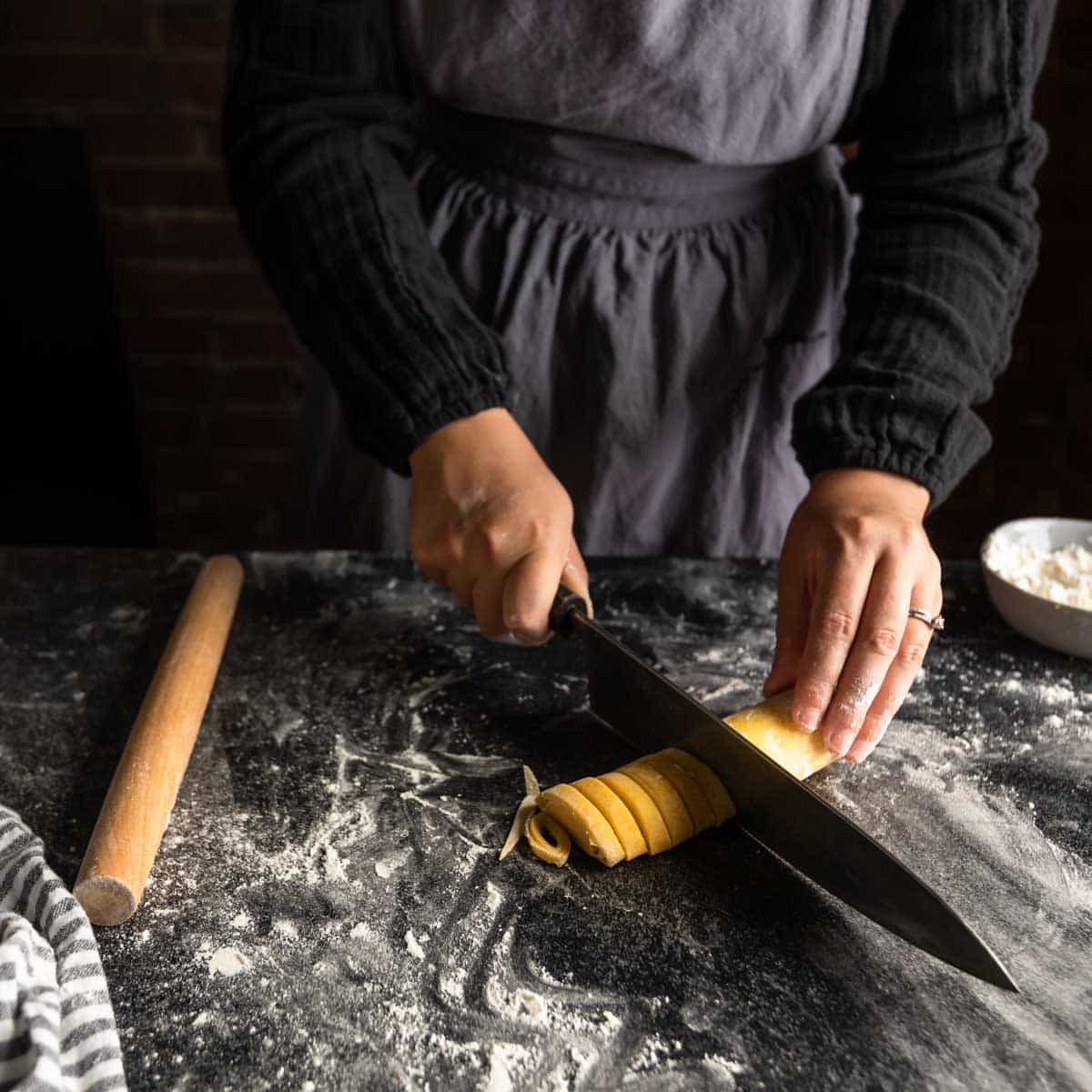 A woman using a sharp knife to cut a log of rolled pasta dough into spirals of homemade fettuccine pasta.