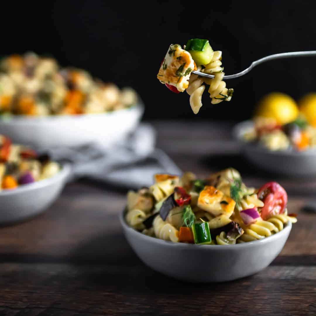 A fork holding a bite of halloumi pasta salad over a small filled dish.