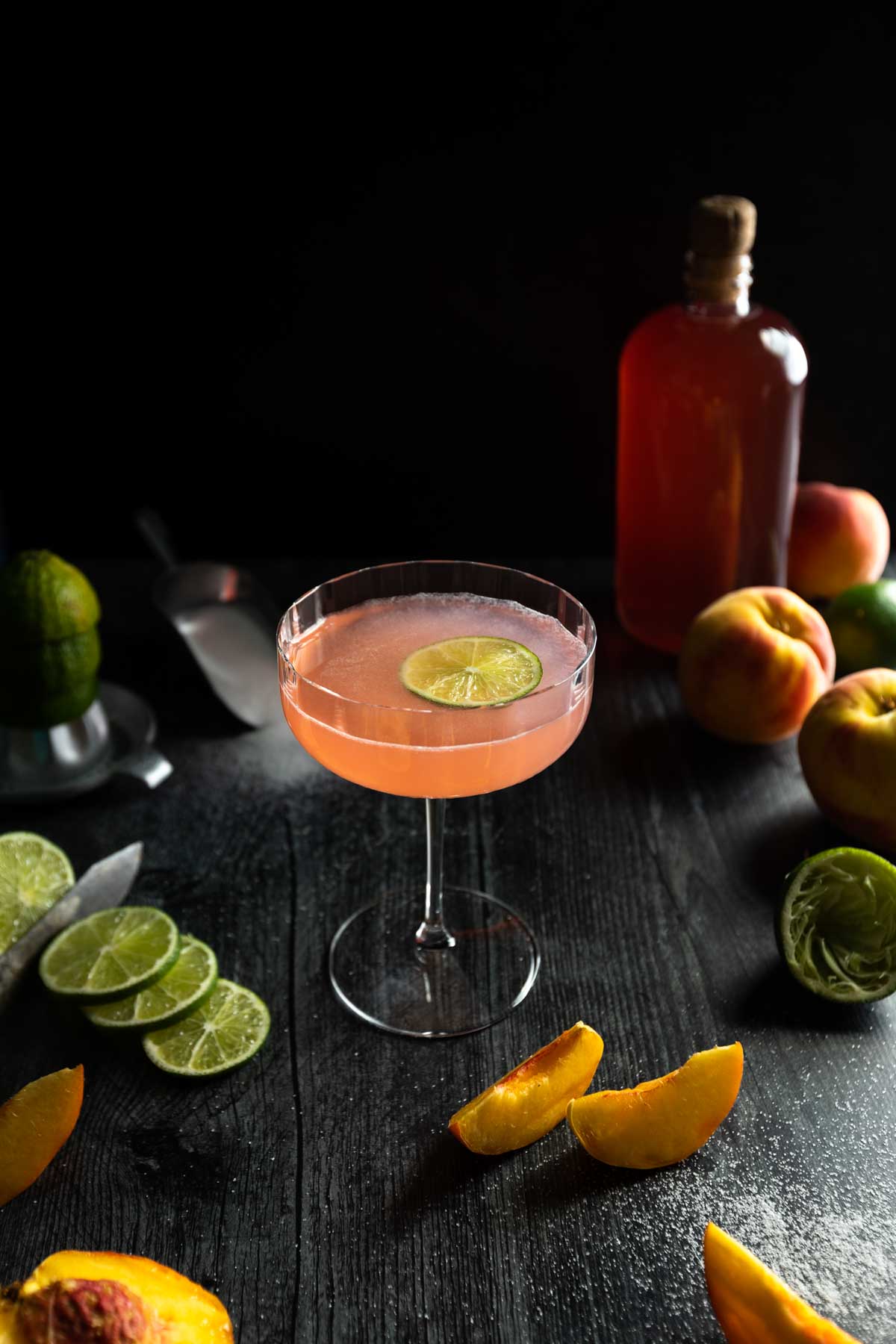 A pink peach daiquiri in a fluted coupe glass with a lime wedge garnish surrounded by slices of peaches, limes and a bottle of peach syrup