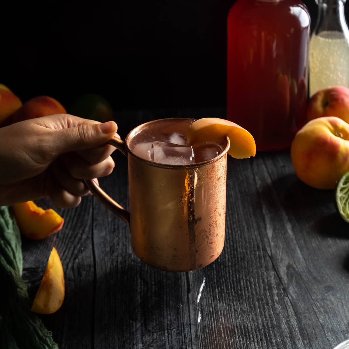 A hand picking up an icy copper mug of Peach Moscow Mule garnished with a fresh peach slice